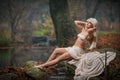 Lovely young lady sitting near river in enchanted woods. Sensual blonde with white clothes posing provocatively in autumnal park. Royalty Free Stock Photo