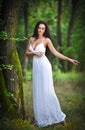 Lovely young lady wearing an elegant long white dress enjoying the beams of celestial light on her face in enchanted woods. Long