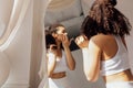 Lovely young mixed race woman in white clothes stands near mirror and reflectes in it