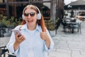 Lovely young woman listening to music through wireless earphones and showing thumbs up gesture to camera on city street Royalty Free Stock Photo