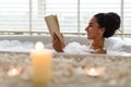 Lovely young woman lying in foamy bath, reading book in relaxing atmosphere with candles, indoors. Copy space Royalty Free Stock Photo