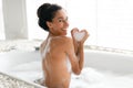 Lovely young woman taking hot bubble bath, making foamy heart with her hands, enjoying spa procedure at home