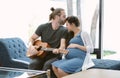 Lovers in a living room. Portrait of handsome hipster husband kissing his smiling pregnant wife and playing guitar sitting on Royalty Free Stock Photo