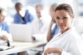 She loves her job. A beautiful medical professional looks at the camera with colleagues working in the background.