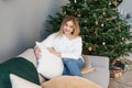 Lovey woman is sitting on a gray sofa near the Christmas tree in the living room and holding a soft pillow in her hands and