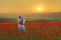 Loving Couple Hug One Another During Romantic Date In Marvellous Spring Poppy Field With Bright Sunset Above Forest And Mountains
