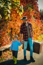 Loving dad carefuly holds hand of his little son. Bearded father wearing hipster outfit and holding large vintage travel