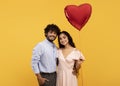 Loving indian couple holding big red heart shaped balloon, hugging and smiling at camera over yellow background