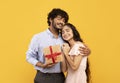 Loving indian man holding wrapped gift box and embracing happy girlfriend, standing over yellow studio background