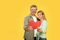 Loving middle aged spouses holding red heart shaped card, celebrating St Valentine's Day, yellow background, copy Royalty Free Stock Photo