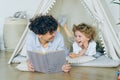 Loving mother reading book to cute child lying on floor in cosy tent indoors