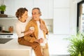 Loving senior married couple enjoying morning coffee at home, happy woman sitting on kitchen table, hugging her husband Royalty Free Stock Photo