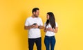 Loving smiling young adult couple dressed in white t-shirts holding mobile phones and looking at each other while standing against Royalty Free Stock Photo