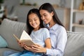 Loving young asian mother reading book with her pretty daughter, sitting together on sofa at home Royalty Free Stock Photo