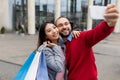 Loving young diverse couple with gift bags taking selfie near huge shopping centre Royalty Free Stock Photo