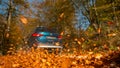 LOW ANGLE: Large blue SUV drives along a road full of brown fallen leaves.
