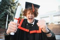 Low angle portrait of happy triumphant male graduate standing near university holding up diploma. From below of young