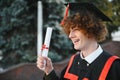 Low angle portrait of happy triumphant male graduate standing near university holding up diploma. From below of young