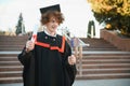 Low angle portrait of happy triumphant male graduate standing near university holding up diploma. From below of young