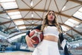 Low-angle shot of a blond cheerleader holding a basketball under her arm and squinting at the camera. Blurred bakground Royalty Free Stock Photo