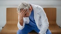 Low angle shot of frustrated, exhausted doctor sitting in hospital corridor. Concept of burnout syndrome among doctors.