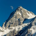 Low angle shot of a rocky mountain peak in snow and the half moon in the blue sky Royalty Free Stock Photo