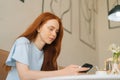 Low-angle view of charming young woman using smartphone texting message and drinking hot coffee sitting at desk in cozy Royalty Free Stock Photo