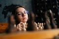 Low-angle view of clever young woman in elegant eyeglasses thinking about chess move sitting on floor in dark room Royalty Free Stock Photo