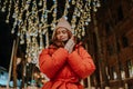 Low-angle view of frozen young woman in hat and winter jacket warming hands in cold winter night standing posing on snow Royalty Free Stock Photo