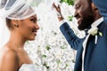 Angle view of happy african american bridegroom touching white veil and smiling near bride and flowers Royalty Free Stock Photo