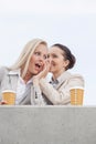 Low angle view of happy businesswomen with disposable coffee cups sharing secrets while standing on terrace against sky