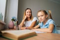 Low-angle view of pretty primary daughter doing homework with young mother sitting at home table by window, looking at Royalty Free Stock Photo