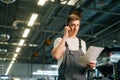 Low-angle view of serious handsome young mechanic male wearing uniform holding clipboard and talking on mobile phone. Royalty Free Stock Photo