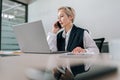 Low-angle view of serious middle-aged businesswoman talking on mobile phone while working typing on laptop sitting at Royalty Free Stock Photo