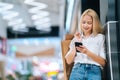 Low-angle view of smiling attractive blonde young woman in stylish clothing using mobile phone holding shopping paper Royalty Free Stock Photo