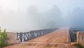 Low cloud in bright mist. single lane road, steel & timber bridge into vanishing forest.