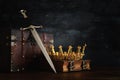 low key image of beautiful queen/king crown over antique book, chest and sword. fantasy medieval period.