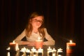 The cute little blonde girl is looking on the light of candle. Lots of candles are around her, over dark background