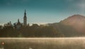 Low profile panoramic photo of lake Bled island with church on a cold hazy foggy early autumn morning. Visible fog coming out of Royalty Free Stock Photo