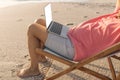 Low section of relaxed senior biracial man sitting with laptop on folding chair at beach