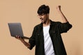 Lucky indian guy using laptop, raising hand up on beige Royalty Free Stock Photo