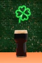 Lucky pub minimal concept. Pint of black stout beer on terracotta table. Shamrock green neon sign above on dark brick background Royalty Free Stock Photo