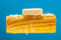 A lump of butter is melted on hot cooked corn. A large ear of yellow corn is shown against a light blue background