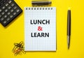 Lunch and learn symbol. Words `Lunch and learn` on white note. Yellow background, paper clips, metallic pen and calculator.