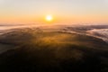 Lush green forest surrounded by farm fields covered by fog with bright orange rising sun. Birseye view of Roztocze