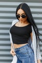 Luxury pretty modern young hipster woman in a summer cape in a black top in jeans with long black hair in stylish sunglasses Royalty Free Stock Photo