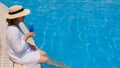 luxury senior woman dangling her legs in an outdoor swimming pool holding a blue refreshing cocktail wearing a straw hat Royalty Free Stock Photo