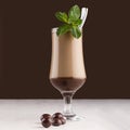 Luxury sweet chocolate milkshake in wineglass with fresh green mint, silver spoon, candies straw on dark brown background, square.