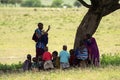 Maasai woman, female teacher teaching young African kids sitting in shade of under Acacia tree in Tanzania, Africa