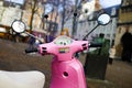 View beyond handlebar, tachometer, windshield and rearview mirrors of retro pink Vespa scooter on blurred city square